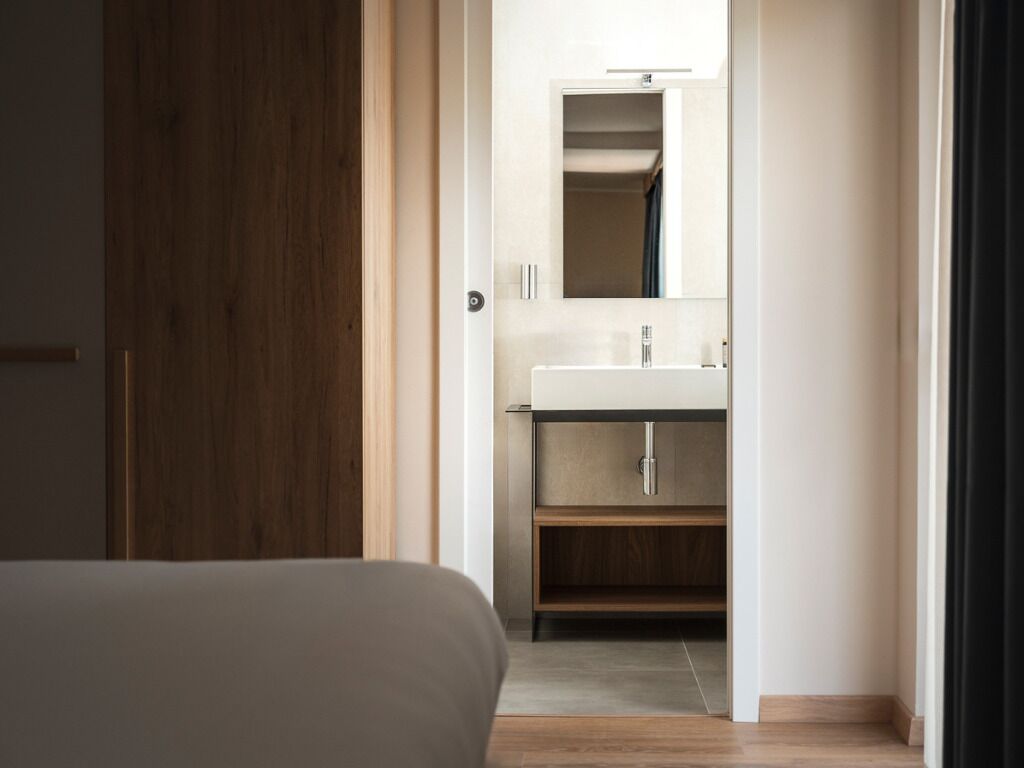 Une chambre avec un plancher en bois, un lit et une salle de bain dotée d’un lavabo et d’un miroir blancs.