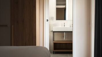 Un dormitorio con piso de madera, una cama y un baño con lavamanos y espejo blancos.