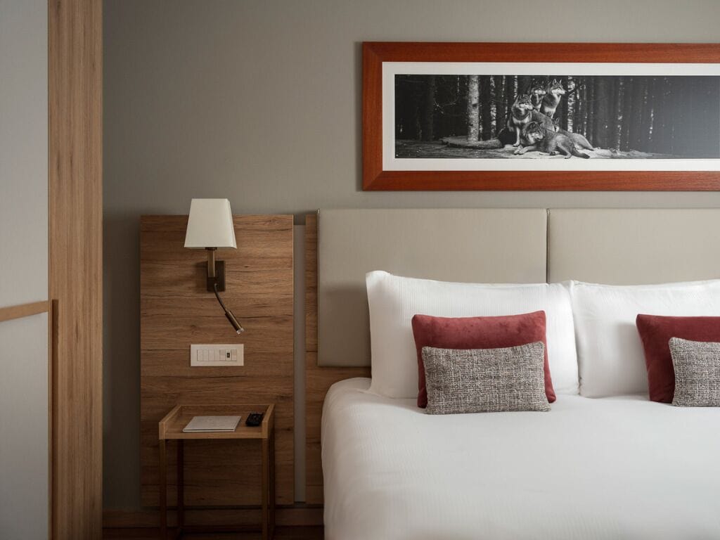 A hotel room with a bed, a nightstand, a lamp, a framed picture on the wall, and a small table with a book.