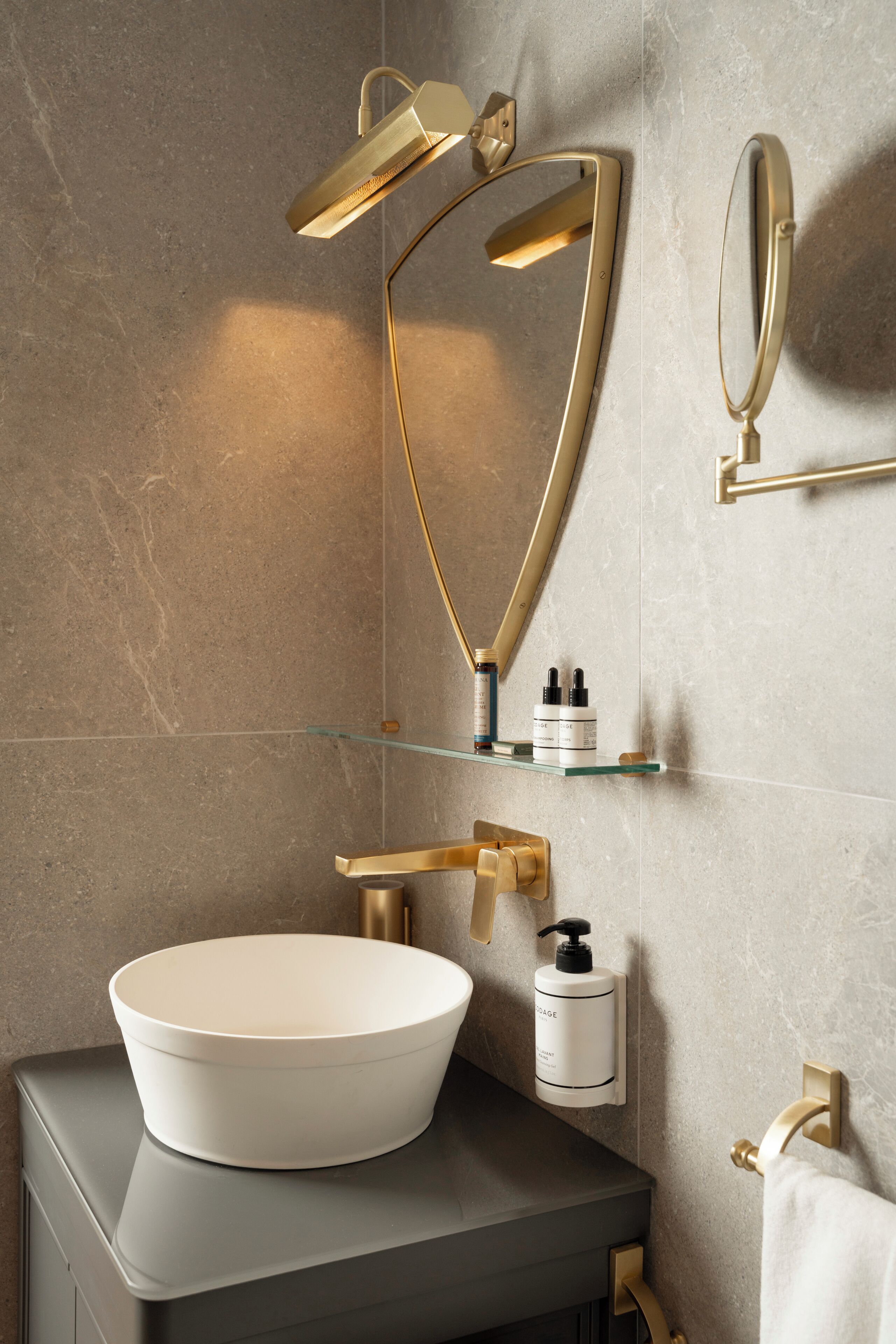 A bathroom with a white vessel sink, gold fixtures, a glass shelf with toiletries, and a wall-mounted mirror.