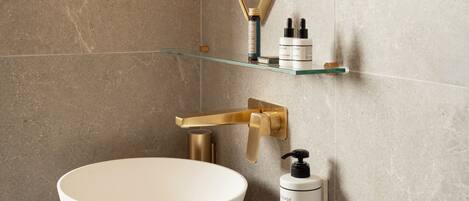 Une salle de bain avec un lavabo sur colonne blanc, des robinetteries dorées, une étagère en verre où sont rangés les articles de toilette et un miroir fixé au mur.