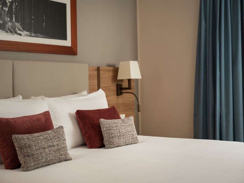 Uma cama bem arrumada com almofadas brancas e vermelhas, uma luminária em uma mesinha de cabeceira e um quadro na parede.