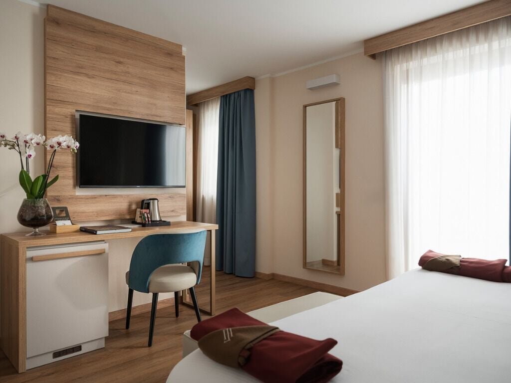 Et hotelværelse med seng, skrivebord med stol, fjernsyn, spejl og et vindue med gardiner.