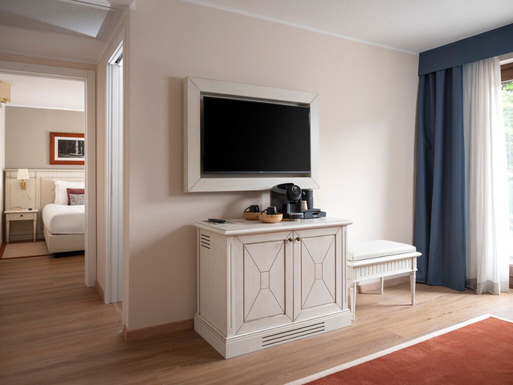 A hotel room with a large flat-screen TV mounted on the wall, a white cabinet with drawers, a bench, and a view of the outdoors through a window with curtains.