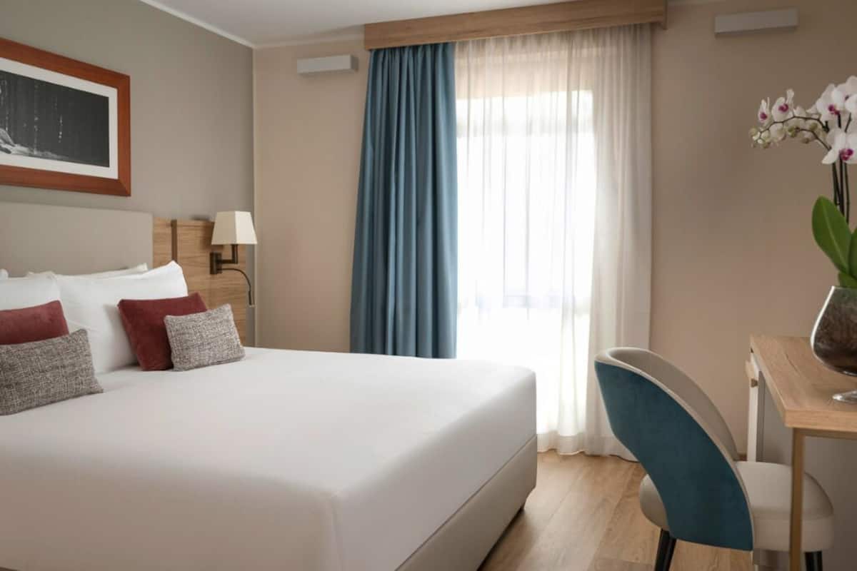Une chambre d’hôtel avec un lit, un bureau, une chaise, une lampe et une fenêtre avec des rideaux.