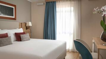 A hotel room with a bed, a desk, a chair, a lamp, and a window with curtains.