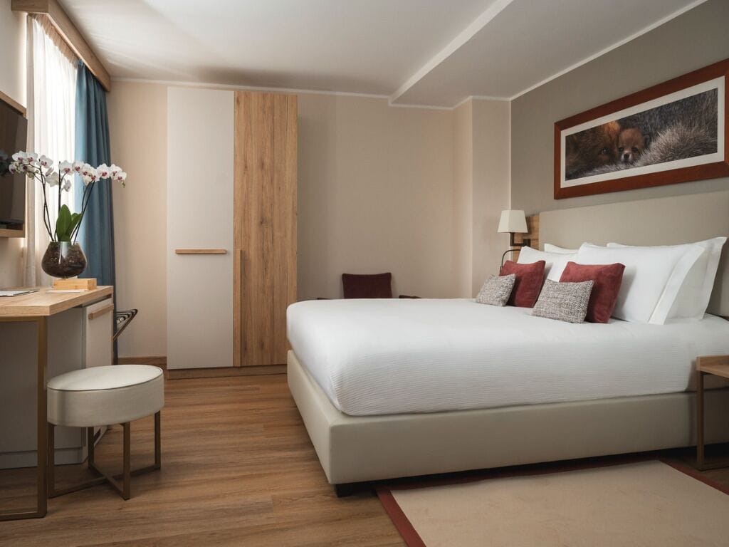A hotel room with a bed, bedside table, lamp, desk, chair, and a window with curtains.