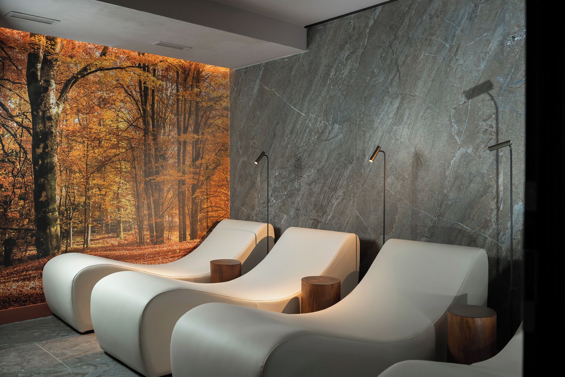 Ein Wellnessraum mit drei weißen Liegestühlen, einem großen Wandgemälde von einem Wald und einer Marmorwand.