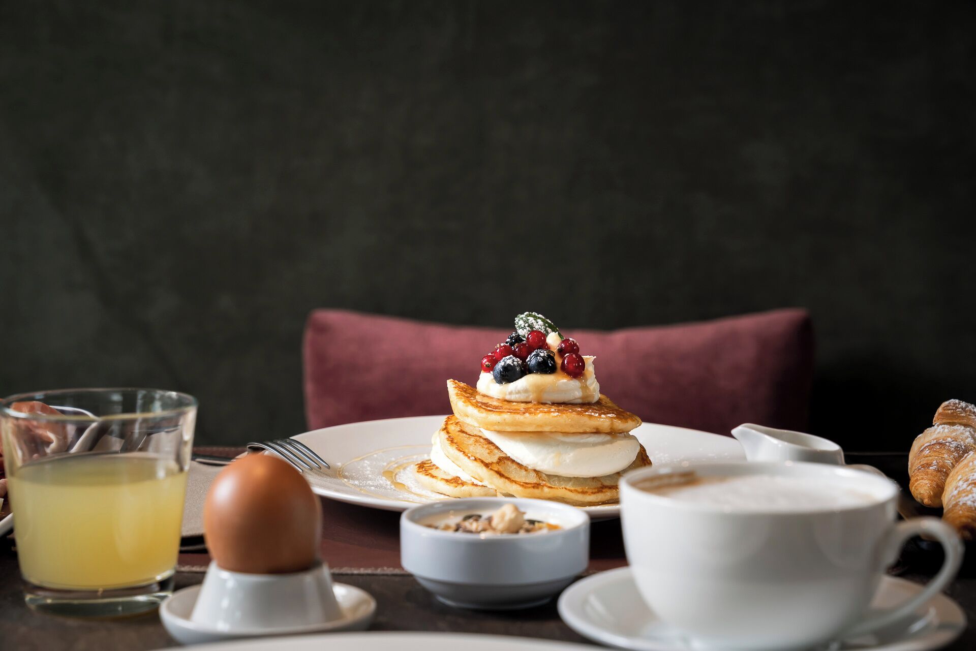 A breakfast setting with pancakes, berries, a croissant, a glass of orange juice, a cup of coffee, and a bowl of cereal.