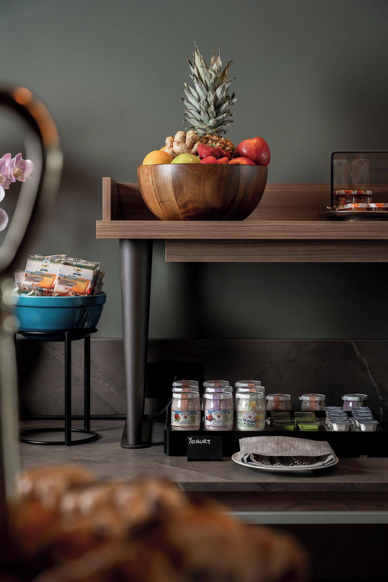 A kitchen counter with a wooden bowl of fruits, a blue bowl of biscotti, and jars of yogurt.