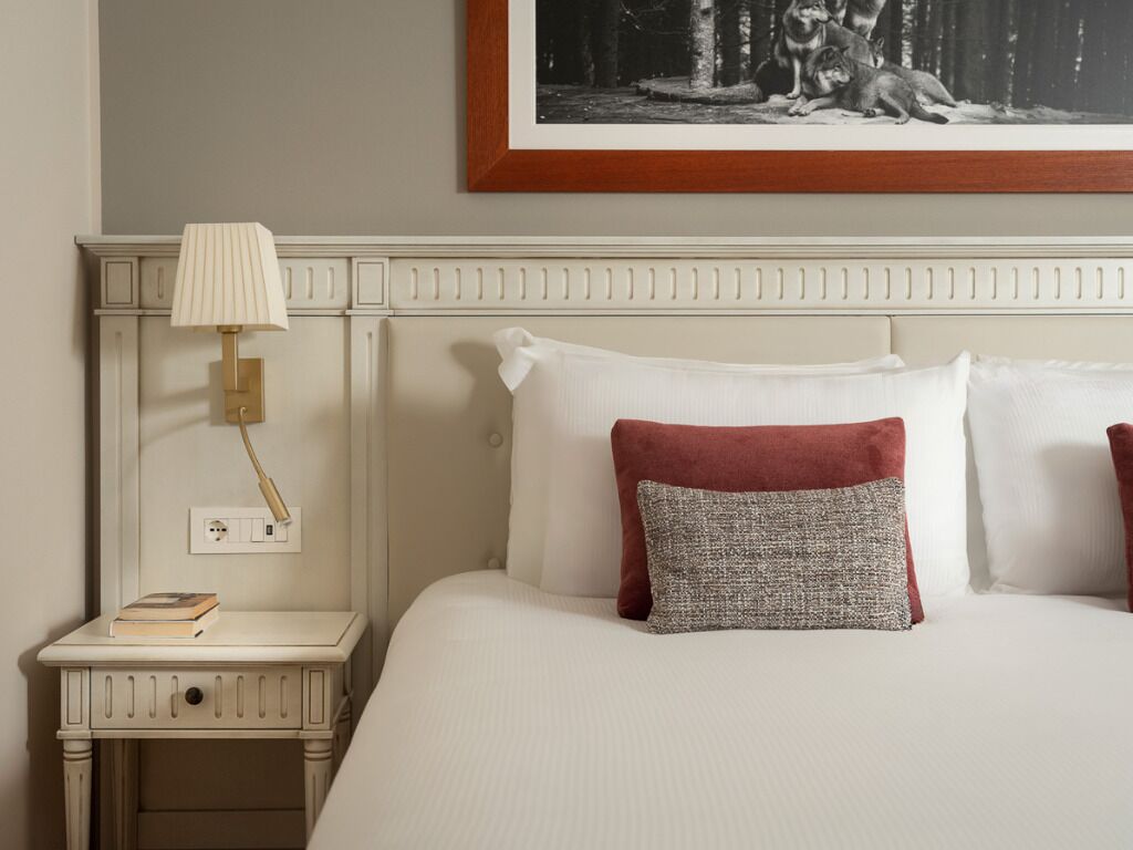 A neatly made bed with white linens and two pillows, one red and one grey, a bedside lamp, and a framed picture above the bed.