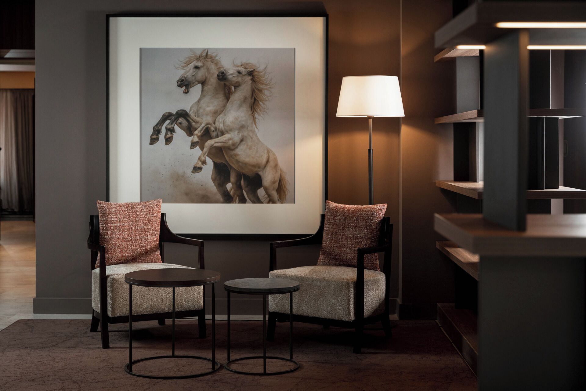 Um hall de hotel moderno com uma obra de arte emoldurada de cavalos, duas poltronas, um abajur e uma estante de design minimalista.