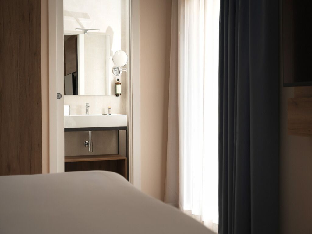 Habitación de hotel con cama, baño con lavabo y espejo, y ventana con cortinas.