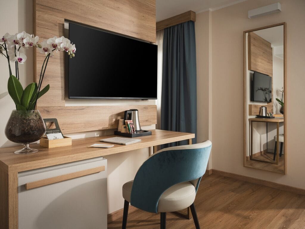 A modern hotel room with a wooden desk, a flat-screen TV, a blue chair, a mirror, and a vase with orchids.