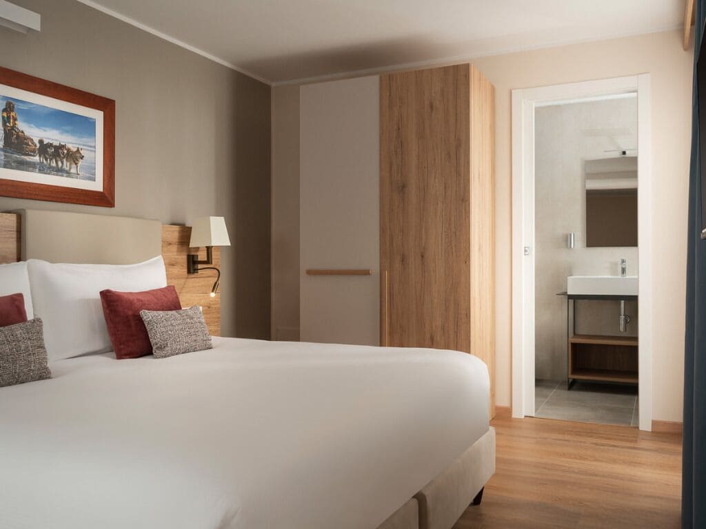 A hotel room with a bed, a nightstand, a lamp, a wooden panel wall, and a bathroom visible through an open door.