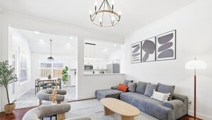 Een moderne woonkamer met een grijze modulaire bank, een houten salontafel en een grijs vloerkleed. Er is een eethoek met een tafel en stoelen, en een keuken op de achtergrond.