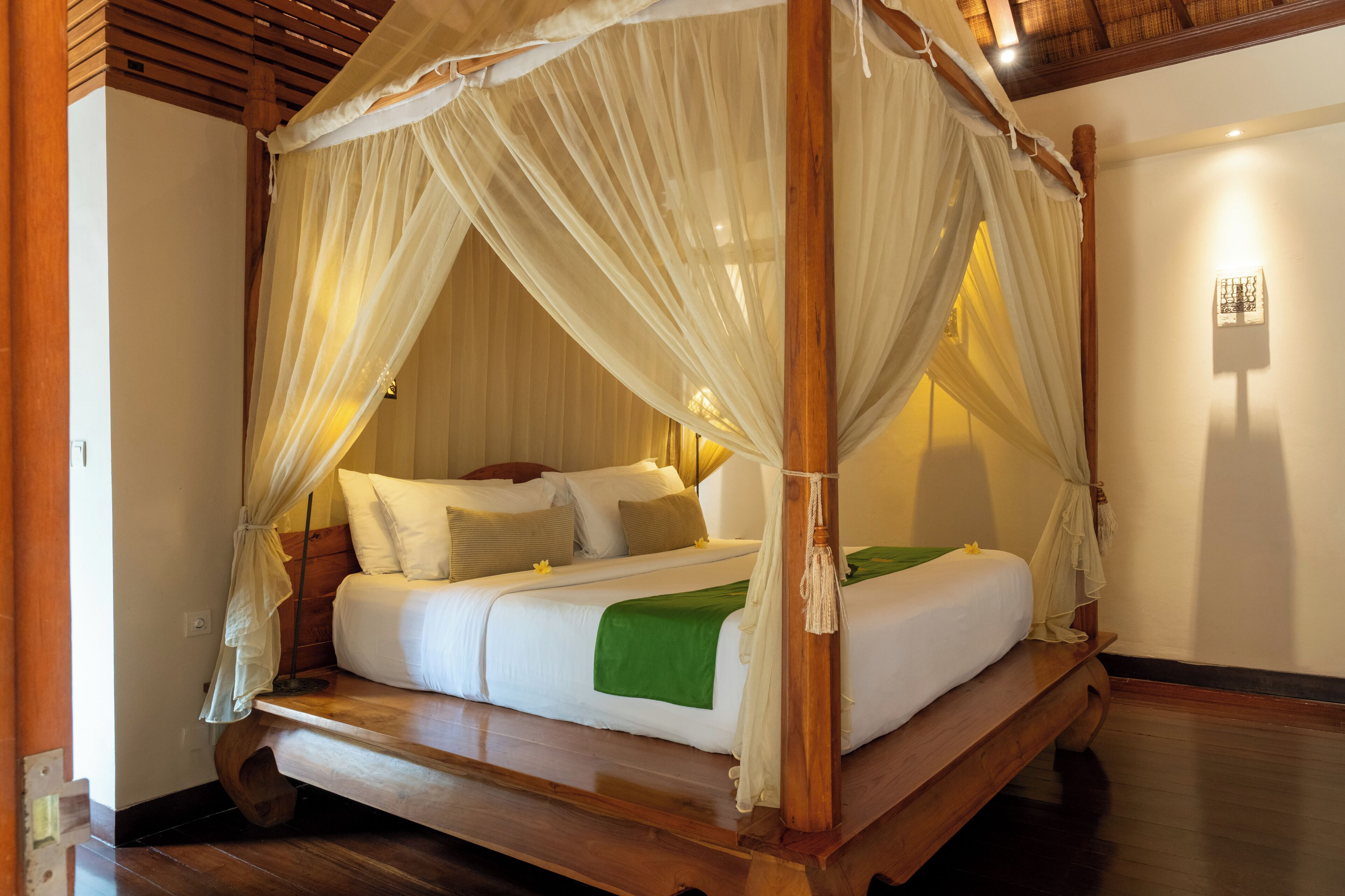 A four-poster canopy bed with white bedding and a green throw blanket, wooden headboard, and a wooden bench at the foot of the bed.