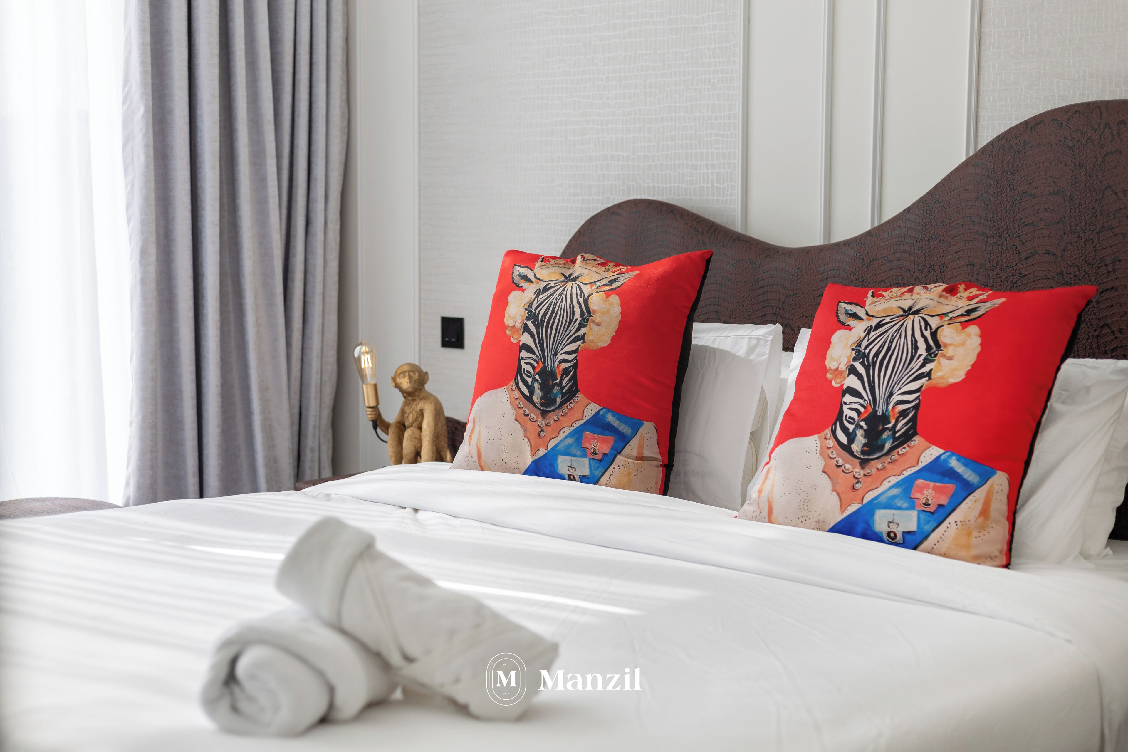A neatly made bed with white linens and two red pillows with a zebra design, a small golden statue, and a window with curtains.