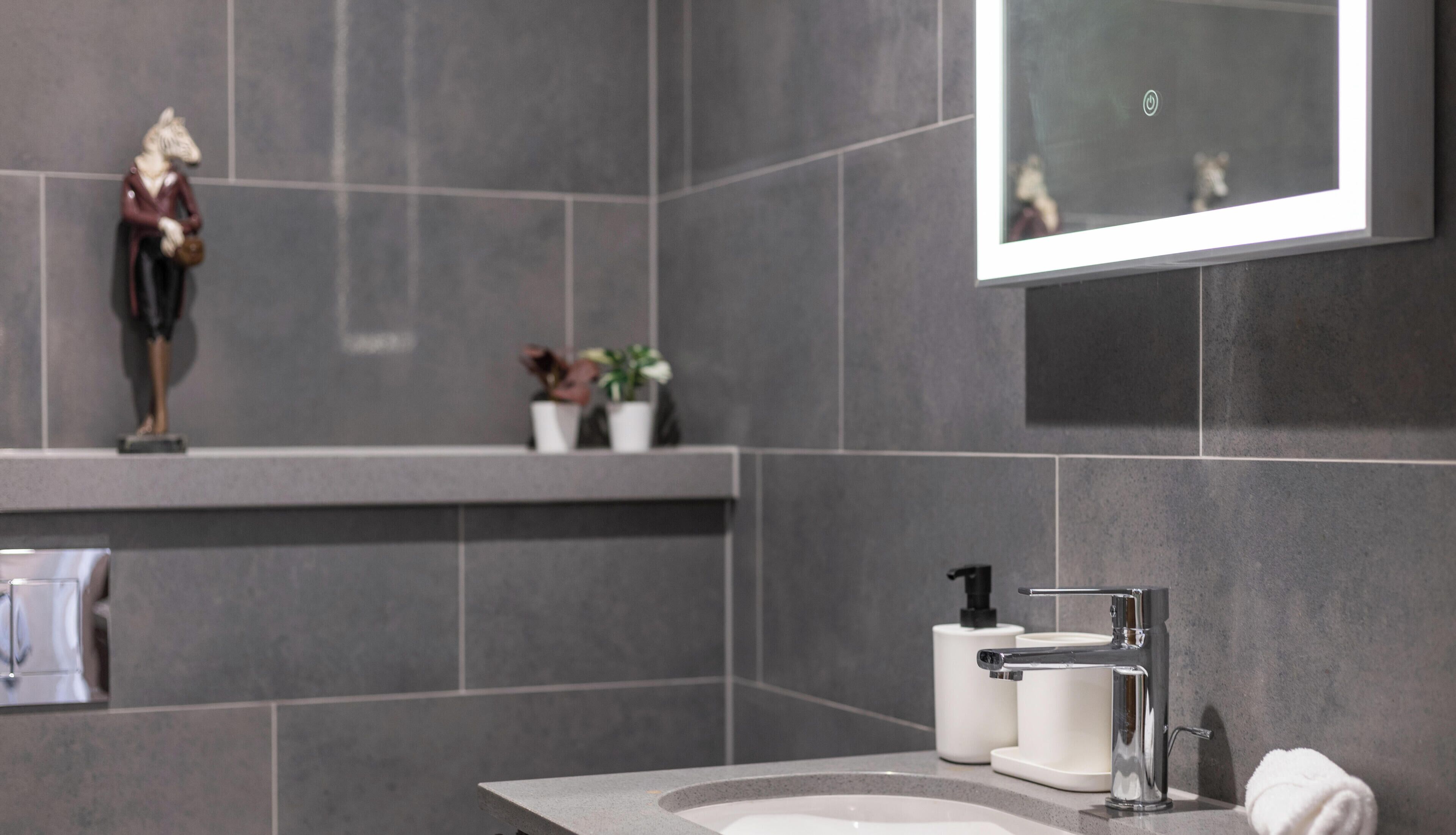 A modern bathroom with a grey tiled wall, a white sink, a mirror, and a towel on the counter.