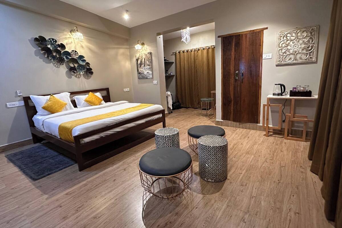 A modern hotel room with a large bed, two round ottomans, a wooden floor, and a small kitchenette area.