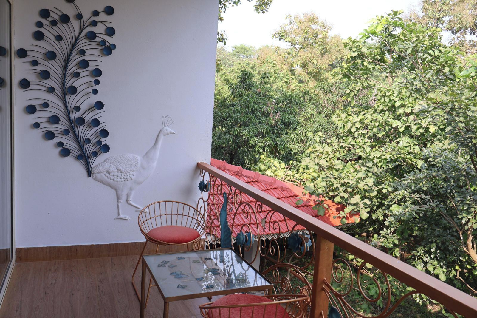 A balcony with a red railing, a white wall with a peacock and circular decorations, a glass table with a red cushion, and lush greenery in the background.