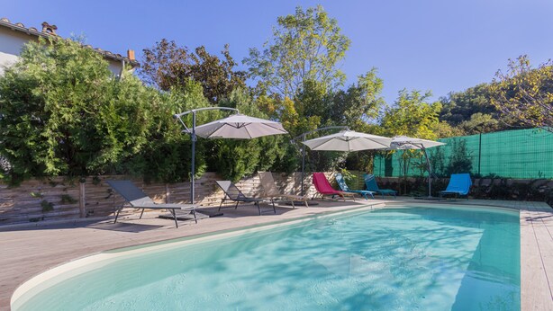 Una piscina con lettini e ombrelloni, circondata da alberi e un pontile in legno.