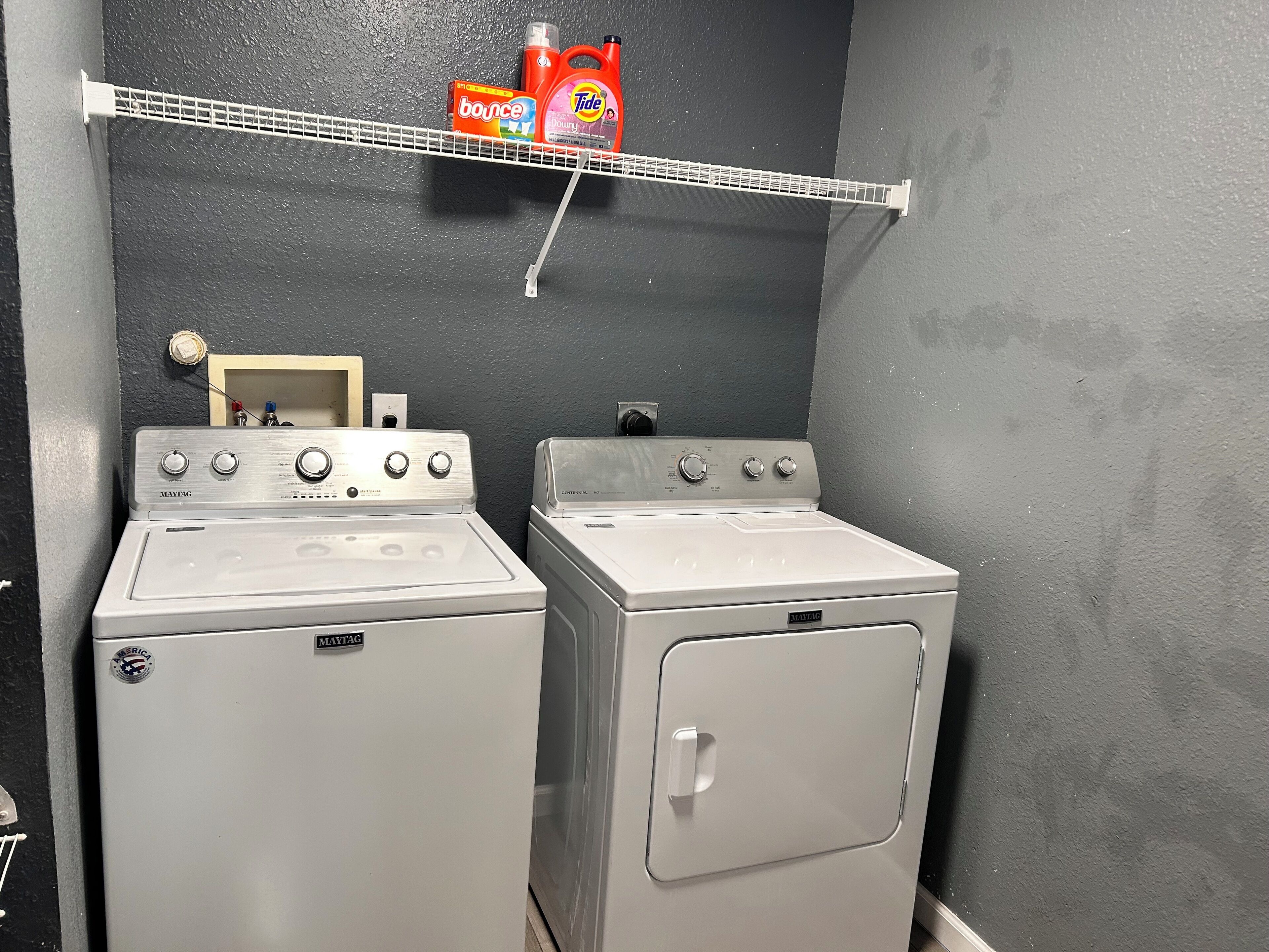 Een wasruimte met twee Maytag wasmachines en drogers, een plank met wasmiddel en een stopcontact.