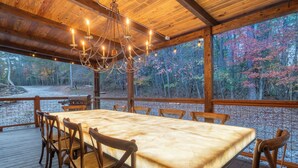 A wooden deck with a table and chairs, a chandelier, and a view of trees.