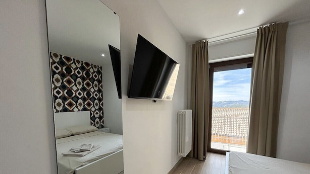 A modern hotel room with a large bed, a flat-screen TV mounted on the wall, a large mirror, and a window with curtains.