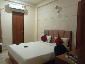 A hotel room with a bed, a desk, a chair, an air conditioning unit, and a door.