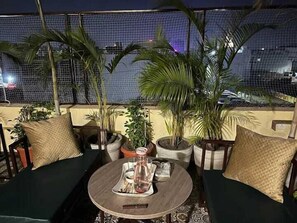 A rooftop terrace with a round wooden table, two cushioned seating areas, and several potted plants.