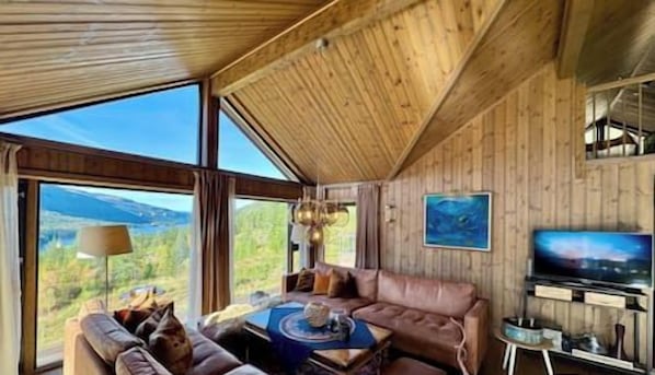 A cabin interior with a large window, wooden walls, a sofa, a coffee table, and a television.