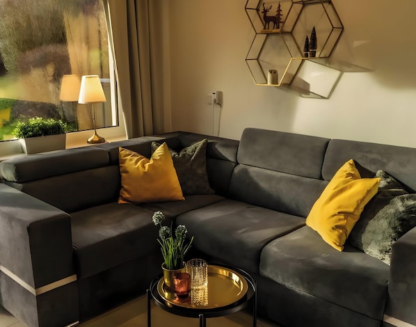 A modern living room with a sectional sofa, a small round coffee table, a potted plant, and a wall-mounted hexagonal shelf with decorative items. - Apartment 'Mellies' with Private Terrace, Garden and Wi-Fi (Willingen (Upland))