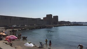 Ein Strand mit einem großen Gebäude im Hintergrund, blauem Himmel und Menschen nahe am Wasser.