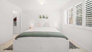 Ein Schlafzimmer mit Bett, Nachttischlampen, einem Fenster mit Jalousien und einer Tür zu einem anderen Raum.