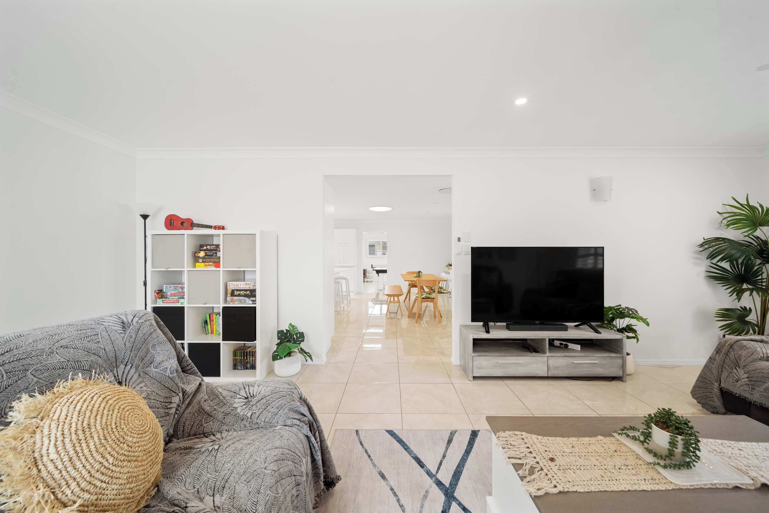 Ein modernes Wohnzimmer mit einem grauen Sofa, einem Couchtisch und einer TV-Konsole. Im Hintergrund ist ein Bücherregal mit verschiedenen Gegenständen und ein Essbereich zu sehen.