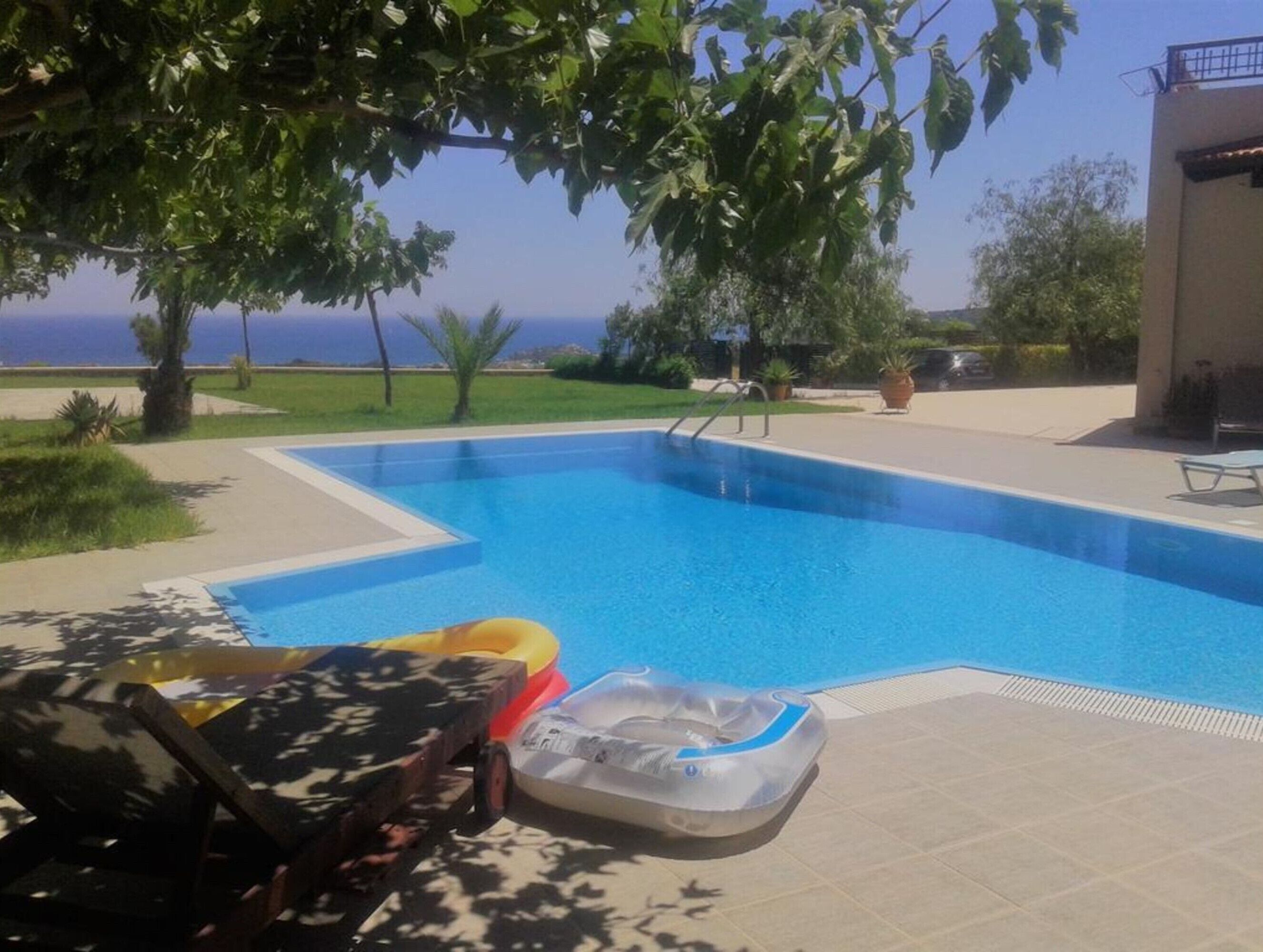 En pool med fri utsikt över havet, omgiven av en uteplats med solstolar och ett träd som ger skugga.
