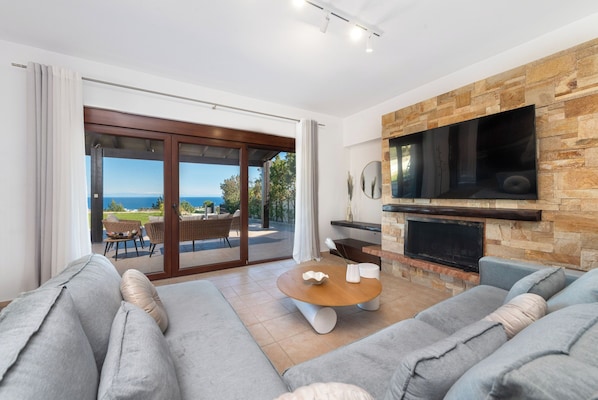 A modern living room with a large TV, stone fireplace, and a view of the sea.