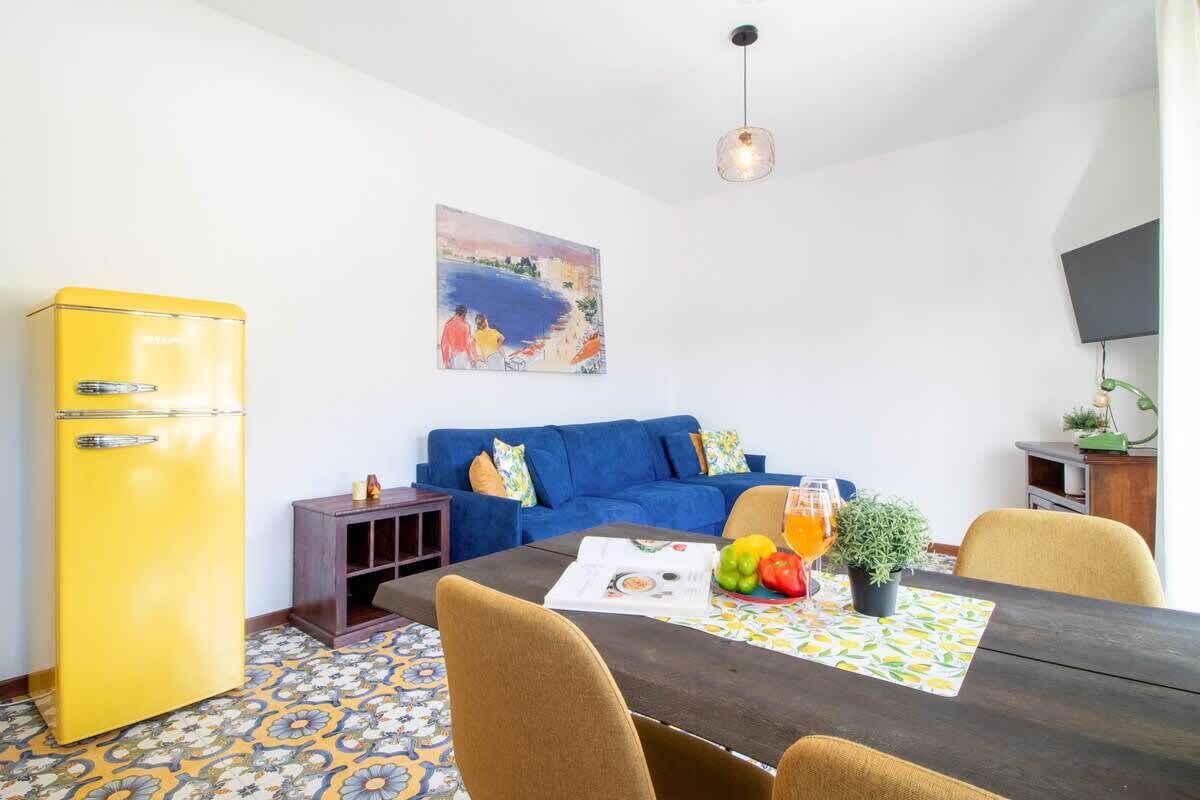 Een ruimtelijk ingerichte leefruimte met een blauwe bank, een gele koelkast, een eettafel met stoelen, en een schilderij aan de muur.