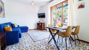 A living room with a blue sofa, a dining table with chairs, a TV mounted on the wall, and a patterned floor.