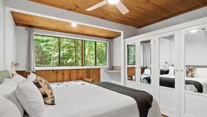 A bedroom with a large bed, wooden ceiling, and a window with a view of trees. - Christmas Creek Rainforest Retreat, ultimate nature filled group getaway. (Lamington)