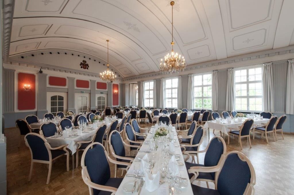Un salón de banquetes con largas mesas dispuestas para comer, candelabros y ventanas amplias.