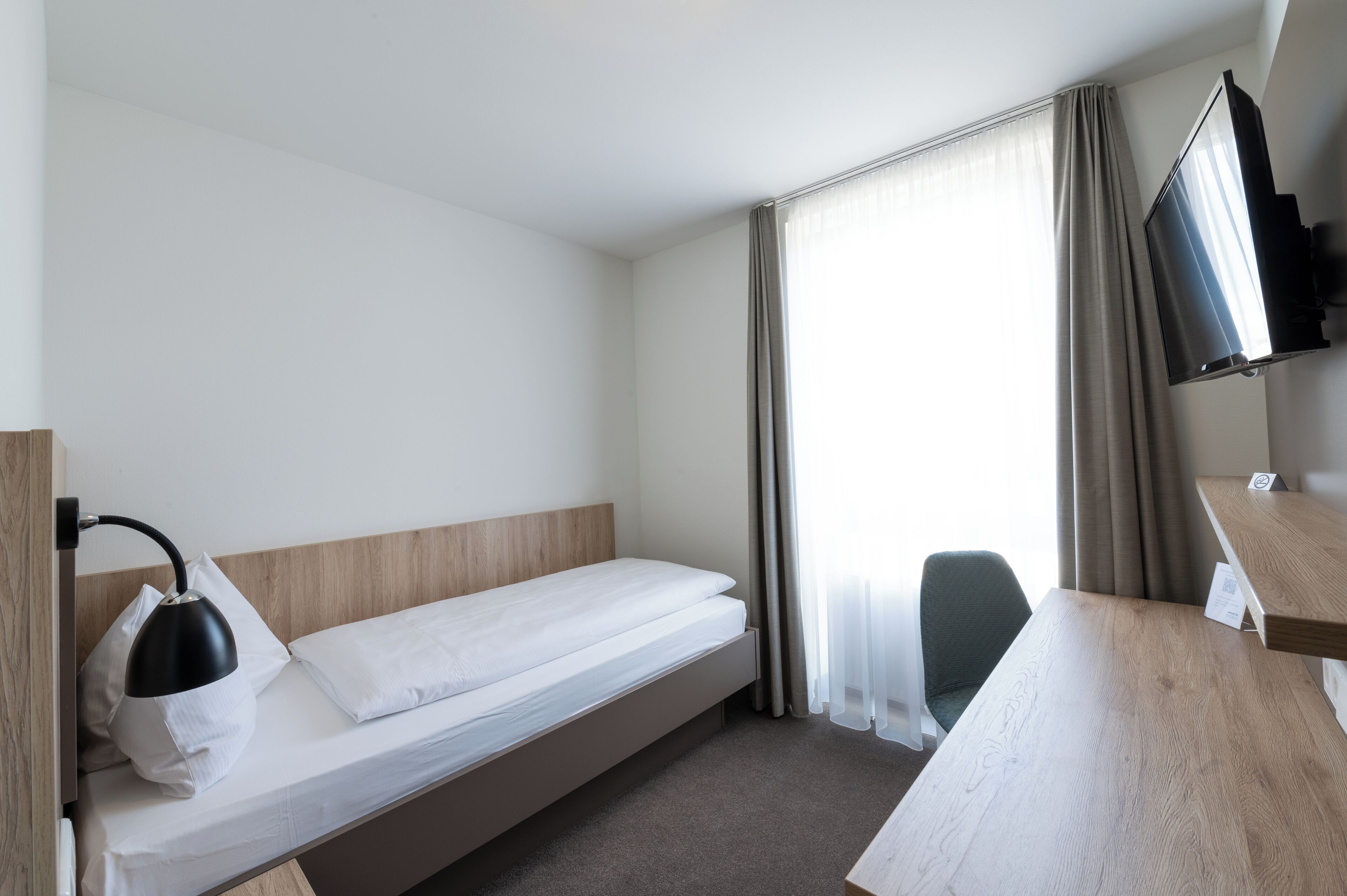 Quarto de hotel com uma cama de solteiro, escrivaninha, cadeira, televisão e janela com cortinas.