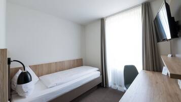 Quarto de hotel com uma cama de solteiro, escrivaninha, cadeira, televisão e janela com cortinas.