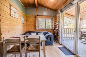 A wooden cabin interior with a dining table, chairs, a sofa, and a sliding glass door leading to an outdoor deck.