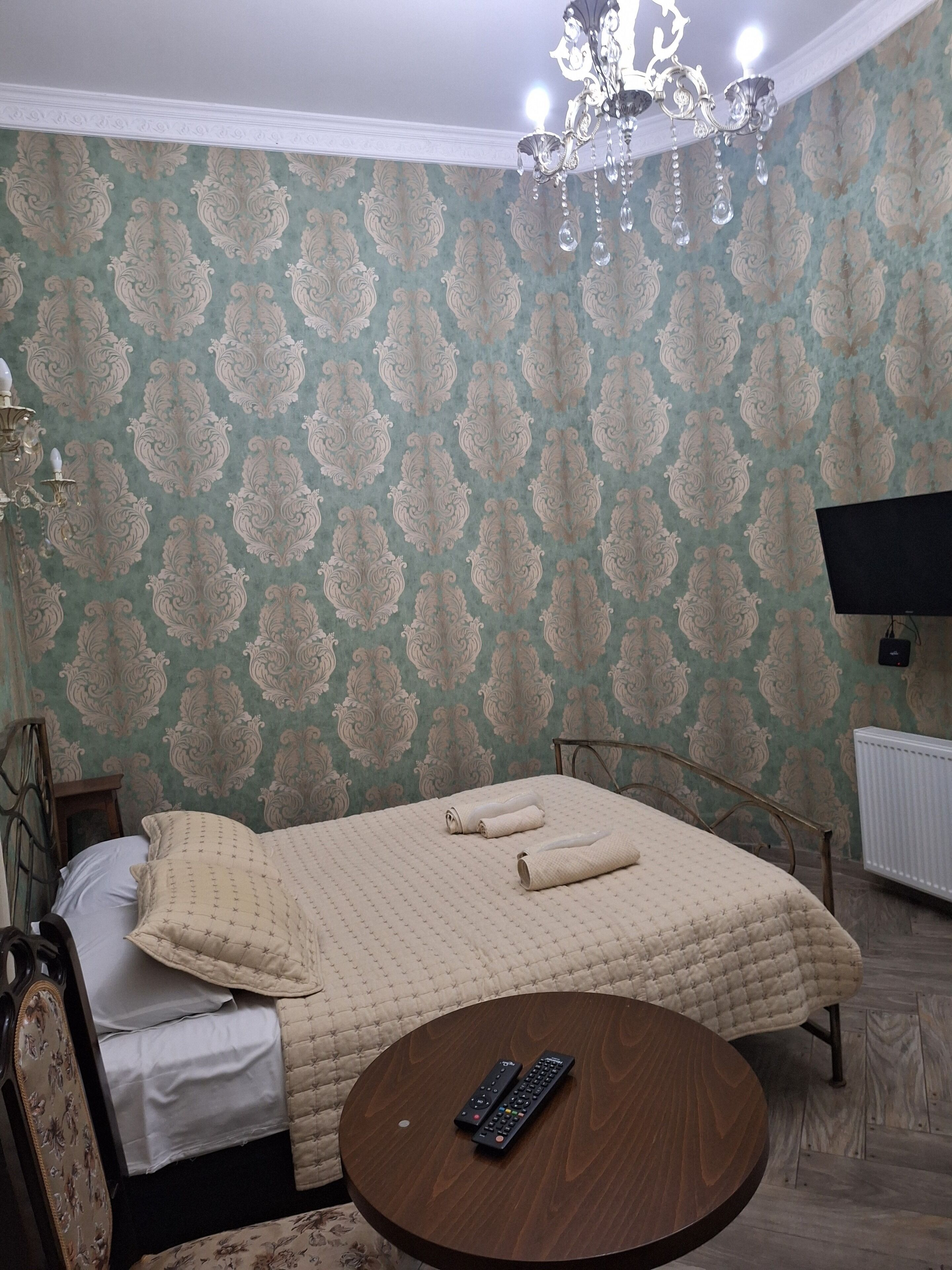 A bedroom with a bed, a round wooden table, a chandelier, and a television on the wall.