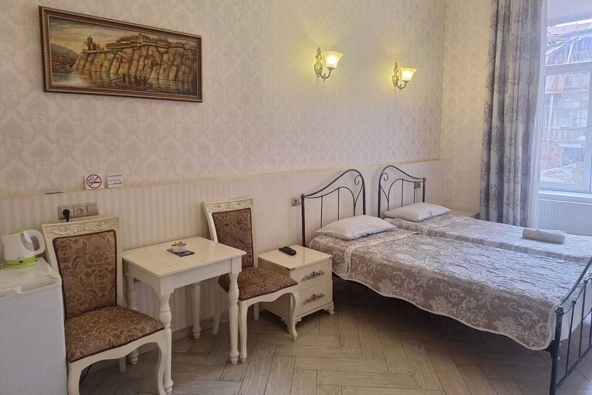 A bedroom with two beds, a desk, and a chair. There is a painting on the wall and two wall-mounted lights.
