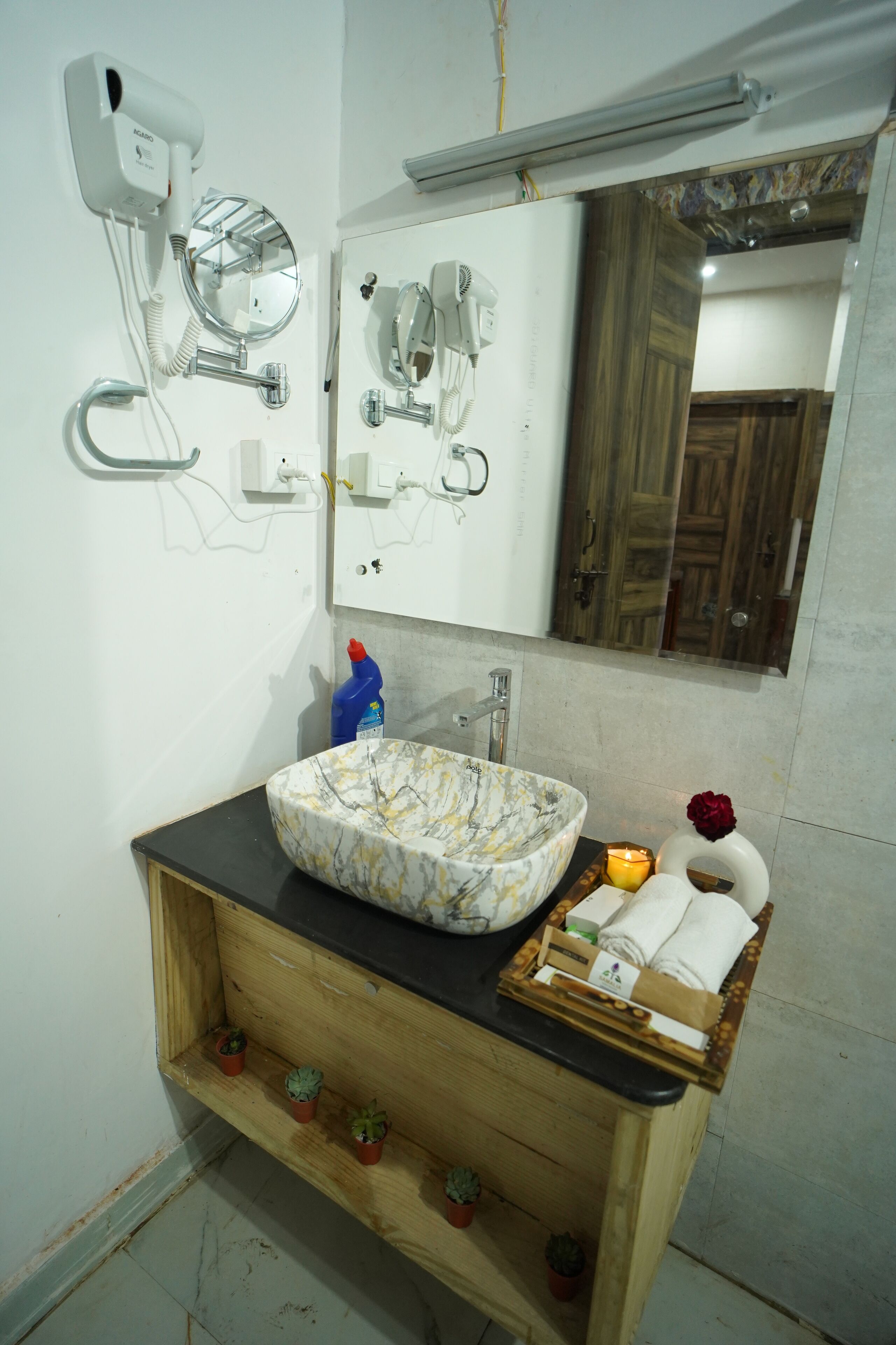 Un baño con lavamanos de mármol, tocador de madera, espejo, secador de pelo y una pequeña estantería con plantas en macetas.