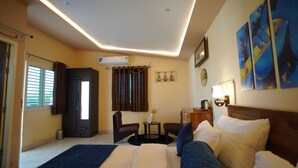 A hotel room with a bed, two chairs, a small table, an air conditioning unit, a window with curtains, and wall art.