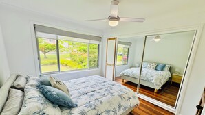 A bedroom with a large bed, a ceiling fan, a window with a view of greenery, and a mirror. - "Bills Place" @ Brunswick - House - Sleeps 8 - Walk to Beach, Creek & Main Town (Brunswick Heads)
